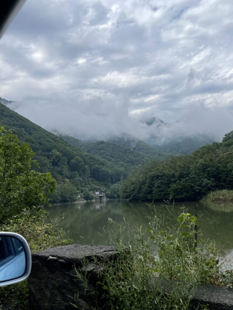 Scenic river valley with forested mountains shrouded in mist, taken from a vehicle viewpoint with a car mirror visible in the foreground
