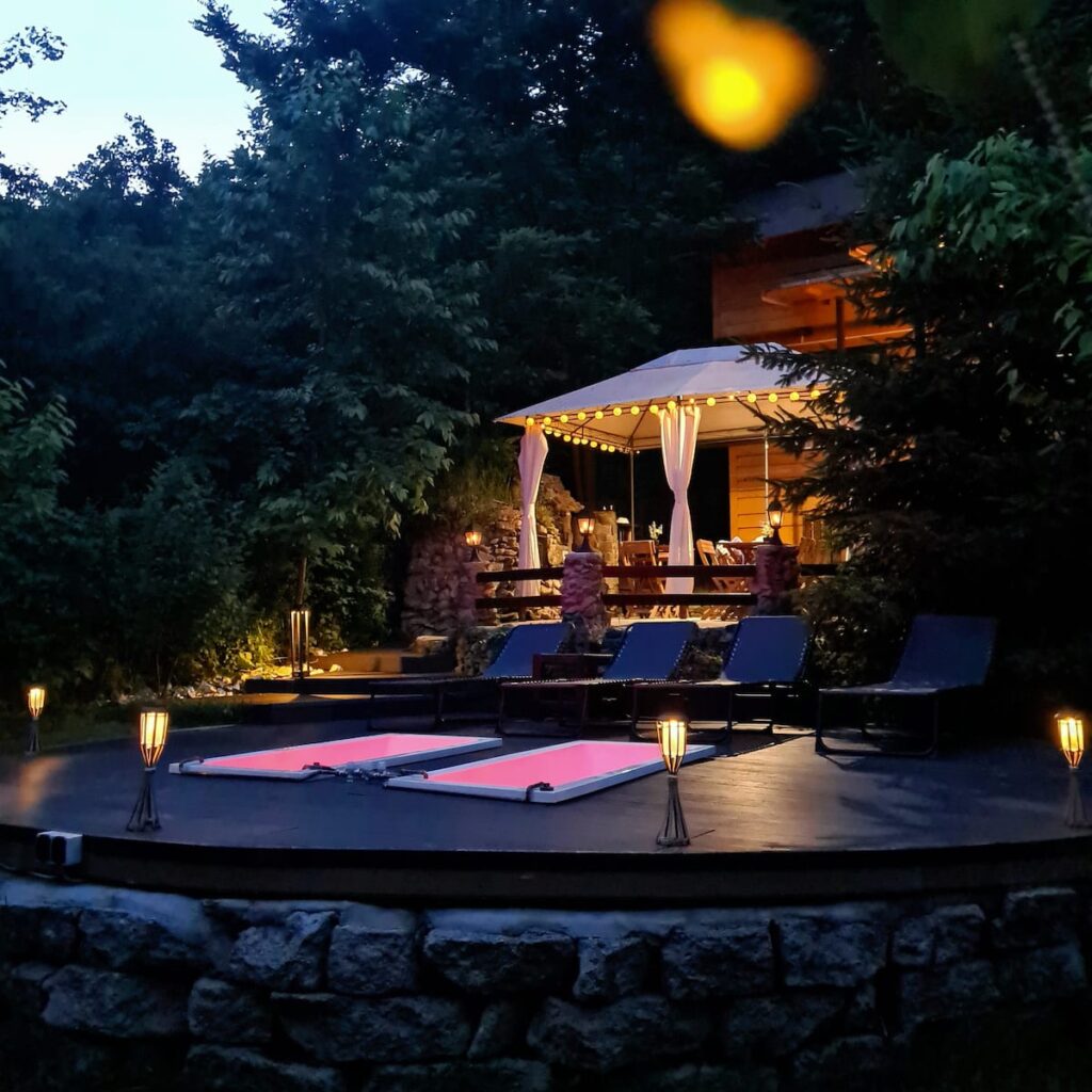 Illuminated backyard patio at dusk with pink yoga mats on a circular platform, lounge chairs, a gazebo with white curtains, lantern lighting, and dense trees surrounding the space