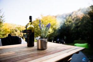 Bottle of red wine and metal mug on wooden table at outdoor camping spot with autumn forest in background