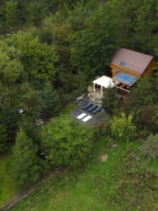 Aerial view of an off-grid cabin in a forest clearing with solar panels on the roof, a white canopy shelter, lounge chairs on a dark patio, and dense green trees surrounding the property