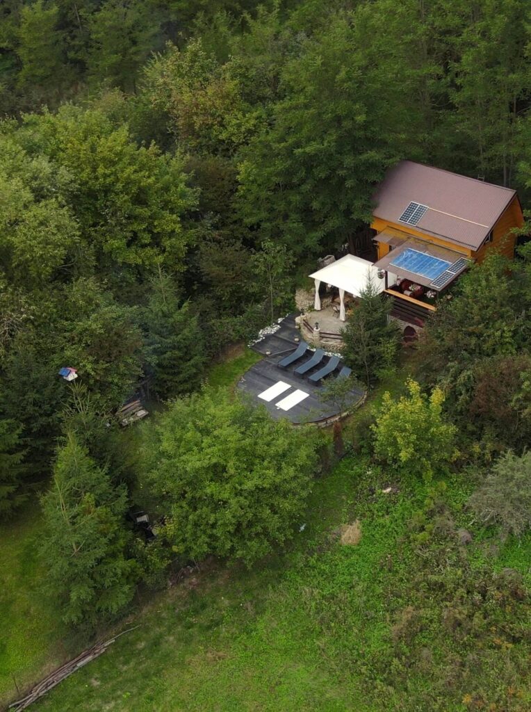 Aerial view of an off-grid cabin in a forest clearing with solar panels on the roof, a white canopy shelter, lounge chairs on a dark patio, and dense green trees surrounding the property