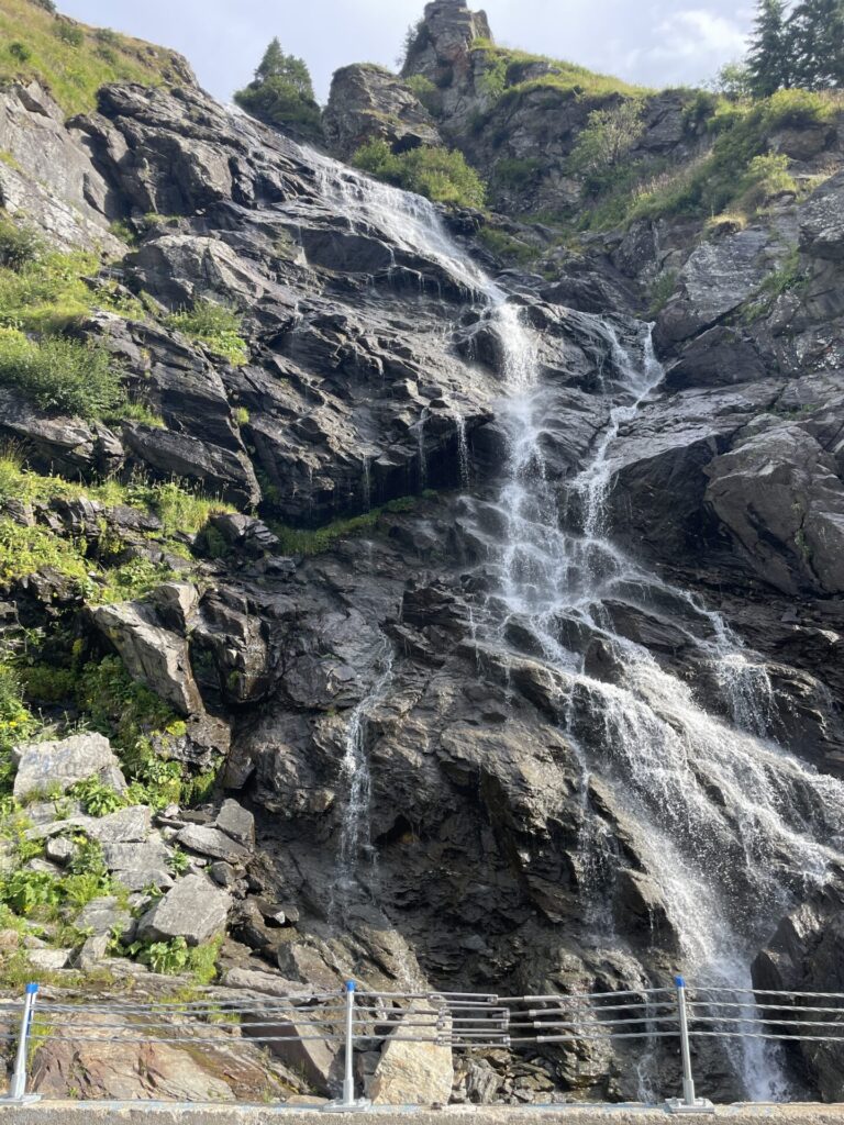 Waterfall cascading down dark layered rock face surrounded by green vegetation, with viewing platform and railing in foreground