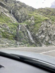 Mountain valley with a waterfall cascading down moss-covered rocky cliffs, viewed from inside a vehicle on a mountain road