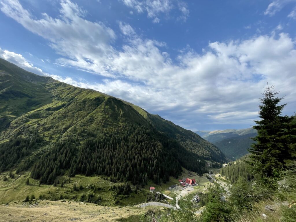 Alpine valley with forested mountainsides, a red-roofed building nestled in the valley floor, and a winding road beneath a blue sky with white clouds
