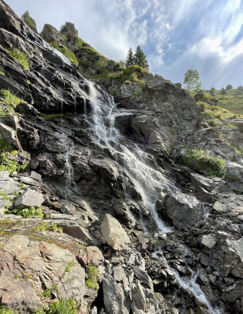 Waterfall cascading down dark layered rock face with moss and vegetation, surrounded by evergreen trees and grassy slopes under a partly cloudy sky