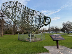 Air Force Heritage Air Park, Winnipeg: Planes, History and Coffee 77 Large green metal satellite dish structure in a park, surrounded by a chain-link fence, with an informational plaque in the foreground and bare trees lining the grassy area