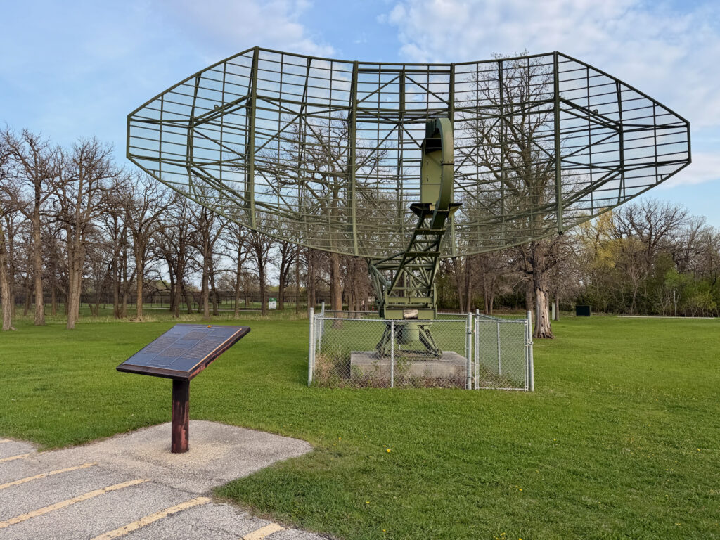 Air Force Heritage Air Park, Winnipeg: Planes, History and Coffee 76 Large green metal radar dish structure mounted on a concrete base with fenced enclosure, displayed in a park setting with interpretive plaque in foreground