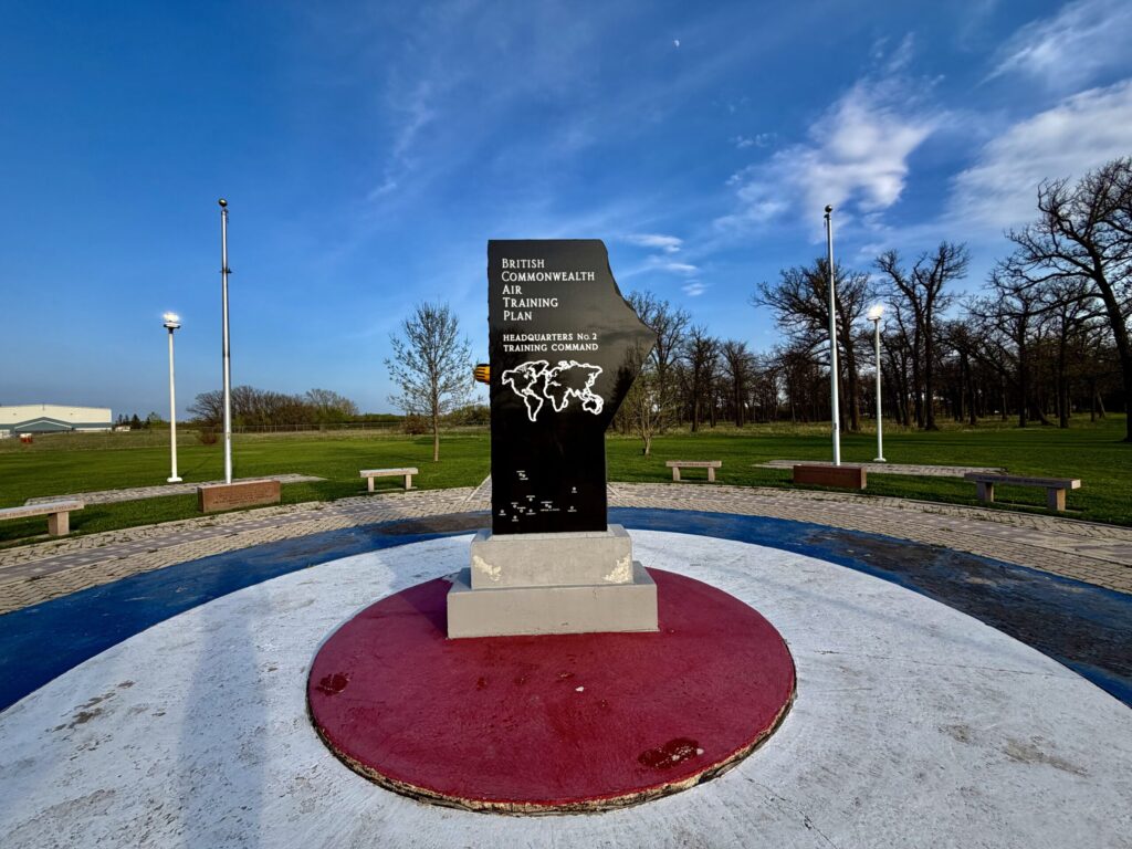 Memorial monument for the British Commonwealth Air Training Plan, Headquarters No. 2 Training Command, featuring a black plaque with world map on a circular base with burgundy and blue sections, surrounded by benches and lamp posts in an open park setting