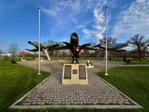 Vintage military aircraft displayed as monuments in a park, with a stone memorial base and plaques in the centre, flanked by two flagpoles under a blue sky