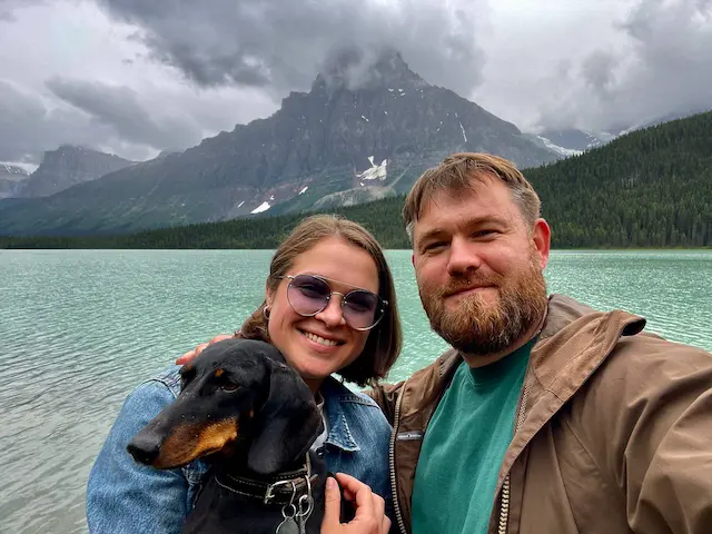 Two people and a black and tan dog pose for a selfie in front of a turquoise alpine lake, with a dramatic jagged mountain peak and forested slopes in the background under cloudy skies