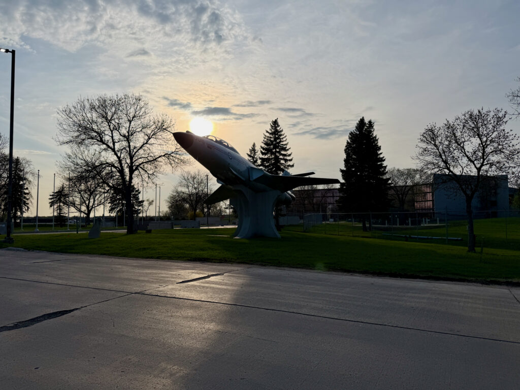 Air Force Heritage Air Park, Winnipeg: Planes, History and Coffee 23 Large fighter jet sculpture mounted on a pedestal in a public park at sunset, with bare trees and evergreens surrounding the installation