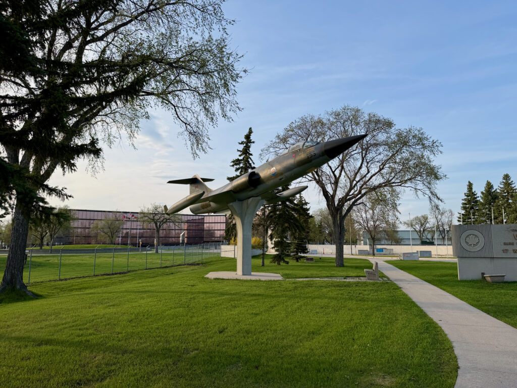 Air Force Heritage Air Park, Winnipeg: Planes, History and Coffee 15 Vintage military jet aircraft mounted on a pedestal in a park setting, surrounded by trees and a concrete pathway, with institutional buildings visible in the background
