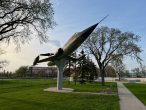 Air Force Heritage Air Park, Winnipeg: Planes, History and Coffee 19 Decommissioned military jet aircraft mounted on a pedestal in a public park, with bare and evergreen trees surrounding a grassy field and sports facilities in the background