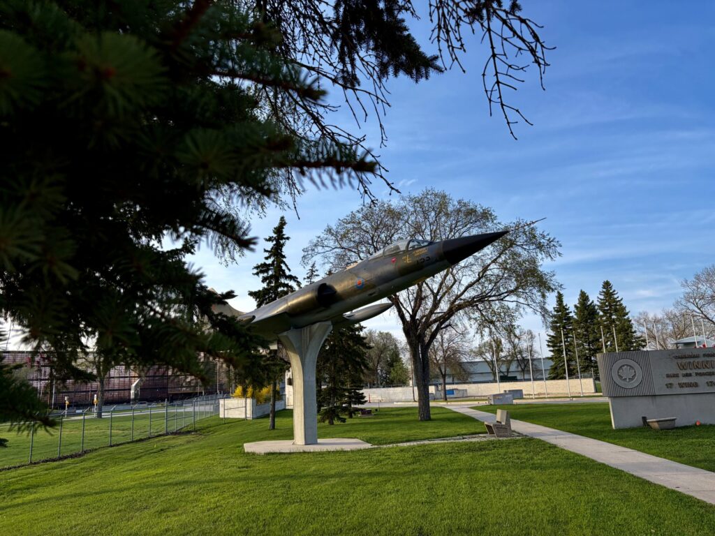 Decommissioned military jet aircraft mounted on a pedestal in a public park, with evergreen trees framing the scene and institutional buildings visible in the background