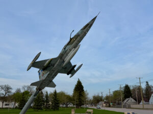 Air Force Heritage Air Park, Winnipeg: Planes, History and Coffee 44 Canadian Armed Forces CF-104 Starfighter jet fighter aircraft mounted on a pole in a public park, with residential buildings and evergreen trees visible in the background
