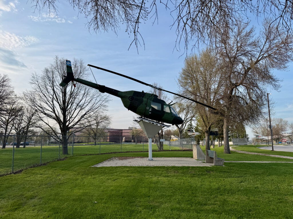 Air Force Heritage Air Park, Winnipeg: Planes, History and Coffee 63 Green military helicopter mounted on a white pedestal in a public park, surrounded by bare and budding trees, with a paved walkway and grassy field