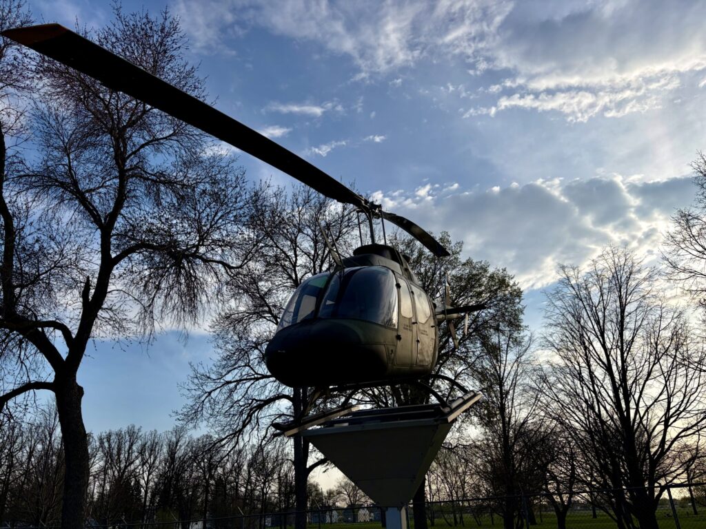 Helicopter suspended from a metal beam structure above a park, surrounded by bare trees and blue sky with clouds