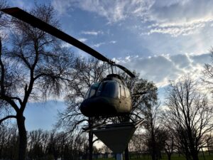 Helicopter suspended from a metal beam structure above a park, surrounded by bare trees and blue sky with clouds