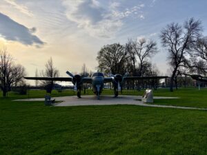 Blue twin-engine aircraft on display in a park with bare and leafy trees, green grass, and an informational marker nearby