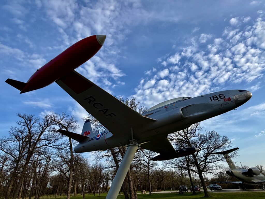 Air Force Heritage Air Park, Winnipeg: Planes, History and Coffee 46 Two vintage military aircraft mounted on display pedestals against a blue sky with white clouds and bare trees in the background