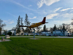 Air Force Heritage Air Park, Winnipeg: Planes, History and Coffee 73 Yellow and burgundy aircraft mounted on a pole in a residential neighbourhood park, with houses and evergreen trees in the background