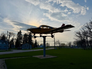 Air Force Heritage Air Park, Winnipeg: Planes, History and Coffee 74 Small aircraft mounted on a pedestal in a public park, silhouetted against a cloudy sky at dusk, with residential buildings and bare trees visible in the background
