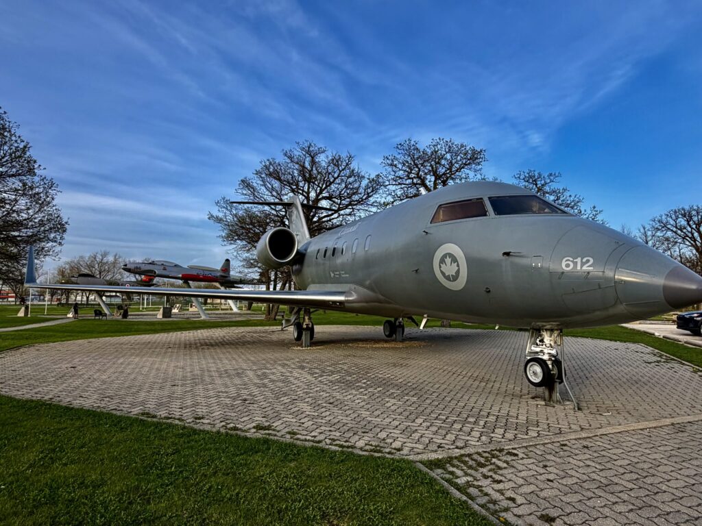 Canadian military transport aircraft on display, marked with number 612 and Canadian Armed Forces insignia, parked on a brick plaza with bare trees and other vintage aircraft visible in the background