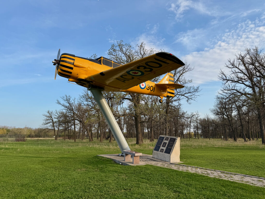 Air Force Heritage Air Park, Winnipeg: Planes, History and Coffee 57 Yellow and black vintage aircraft mounted on a pedestal in a grassy field with bare trees, featuring registration number NC001 and a Canadian military roundel on the fuselage