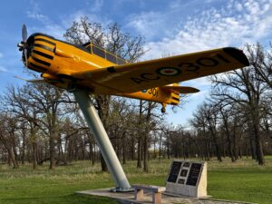 Yellow and black vintage aircraft mounted on a pedestal in a wooded park setting, with registration number visible on the fuselage and a commemorative plaque at its base