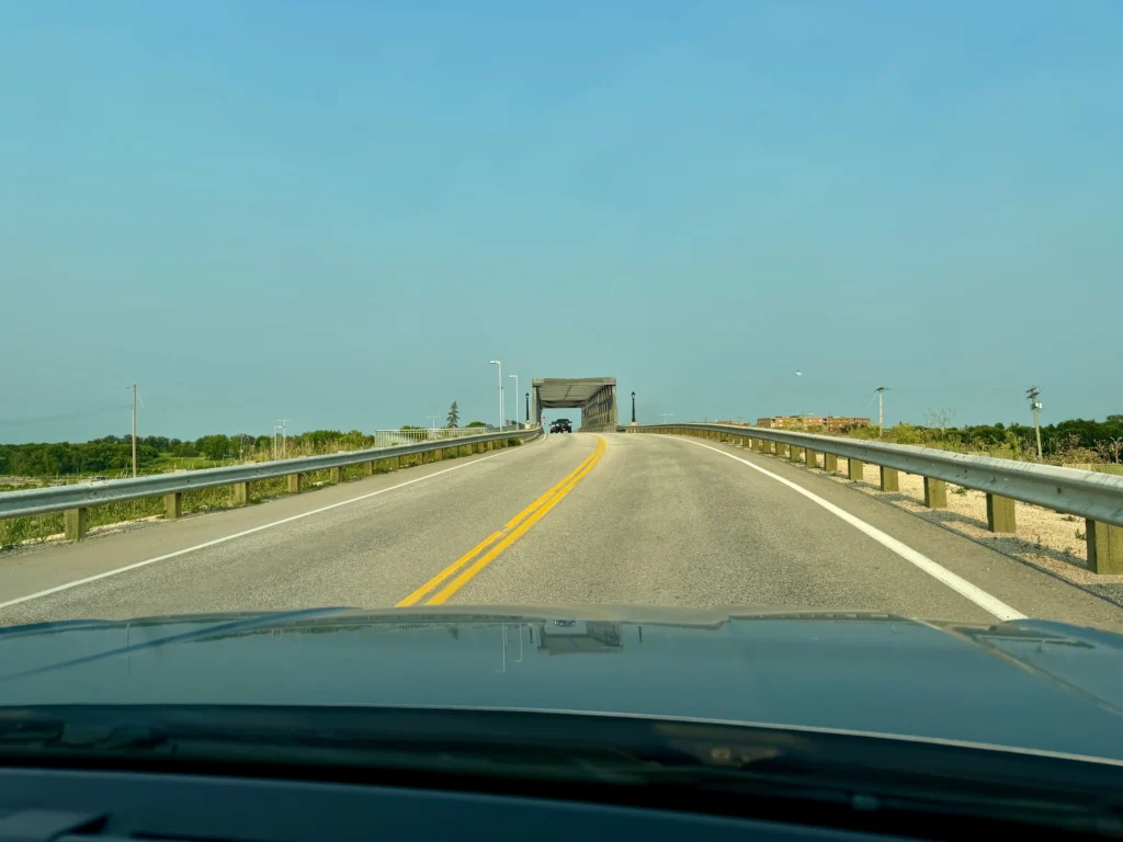 View from inside a vehicle driving across a bridge with yellow centre line markings, metal guardrails on both sides, and a transport truck ahead on the road under clear blue sky