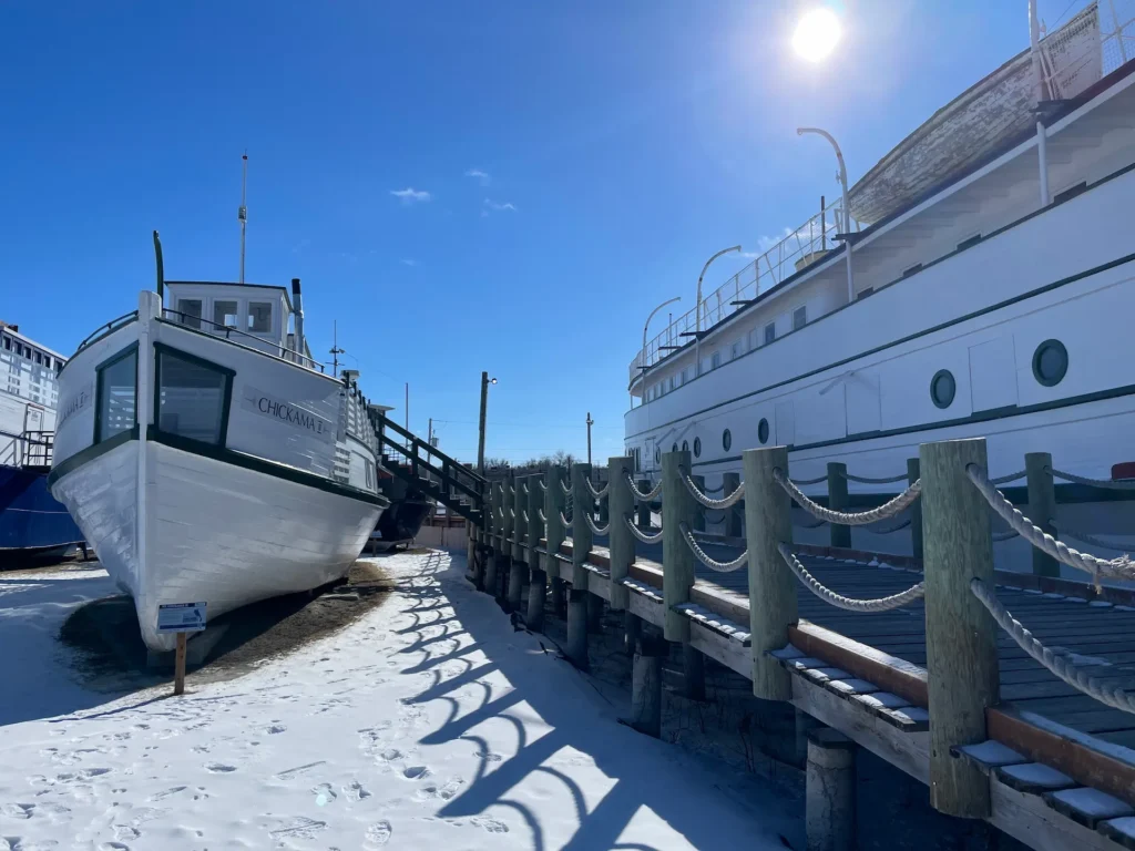 A white passenger ferry named Chickama docked at a wooden pier with rope railings on a sunny day, with a larger cruise ship moored alongside