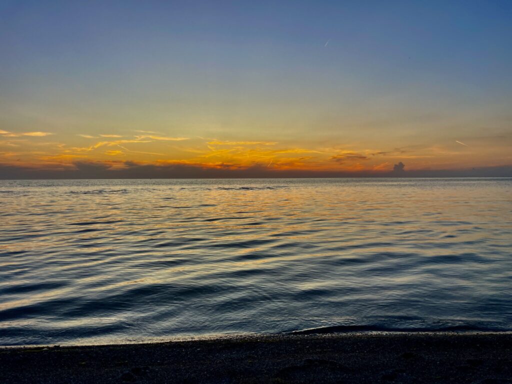 Calm Manitoba lake at sunset with golden and orange sky reflecting on the water, dark pebbled beach in foreground