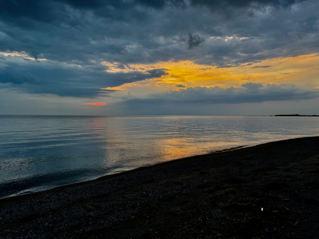Sunset over calm water with dark pebble beach in foreground, dramatic storm clouds lit by golden and orange light breaking through grey sky