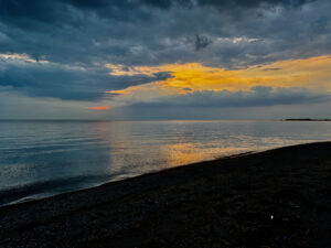 Sunset over calm water with dark pebble beach in foreground, dramatic storm clouds lit by golden and orange light breaking through grey sky
