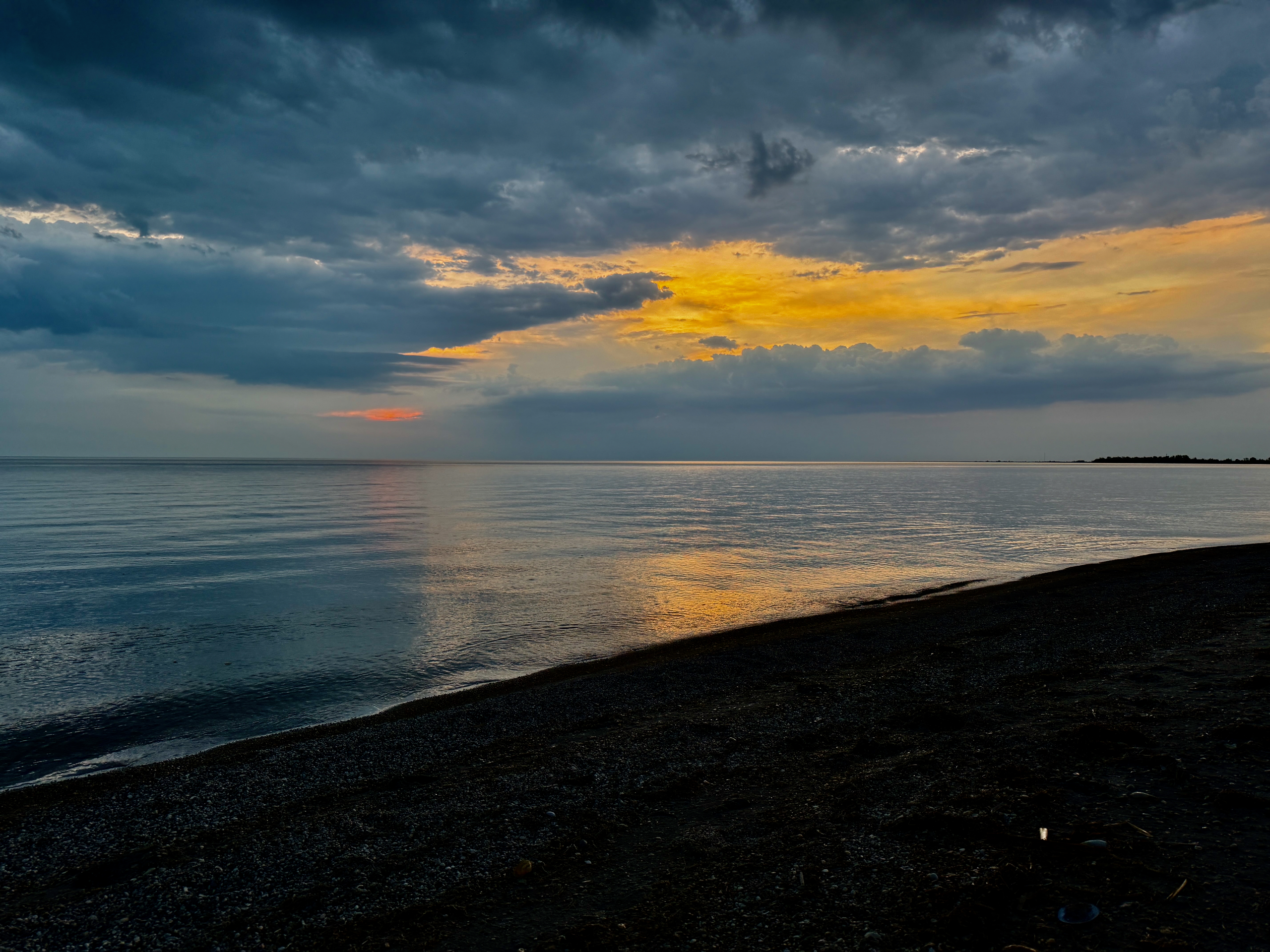 Sunset over calm water with dark pebble beach in foreground, dramatic storm clouds lit by golden and orange light breaking through grey sky