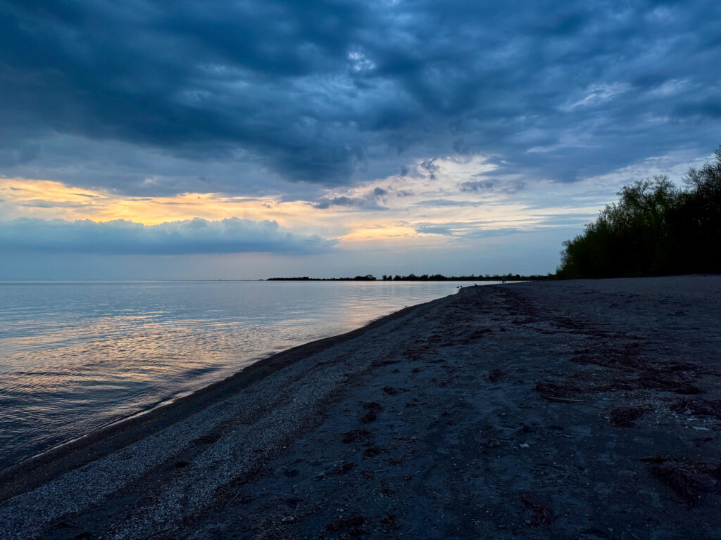 Pebbly beach shoreline at dusk with calm water, distant treeline, and dramatic cloudy sky lit by golden sunset light on the horizon