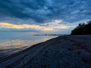 Pebbly beach shoreline at dusk with calm water, distant treeline, and dramatic cloudy sky lit by golden sunset light on the horizon