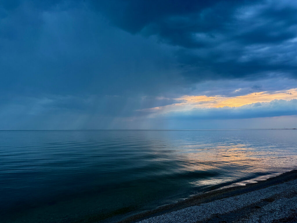 Calm Manitoba lake at dusk with golden light breaking through dark storm clouds on the horizon, pebbled beach in foreground