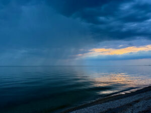 Calm Manitoba lake at dusk with golden light breaking through dark storm clouds on the horizon, pebbled beach in foreground