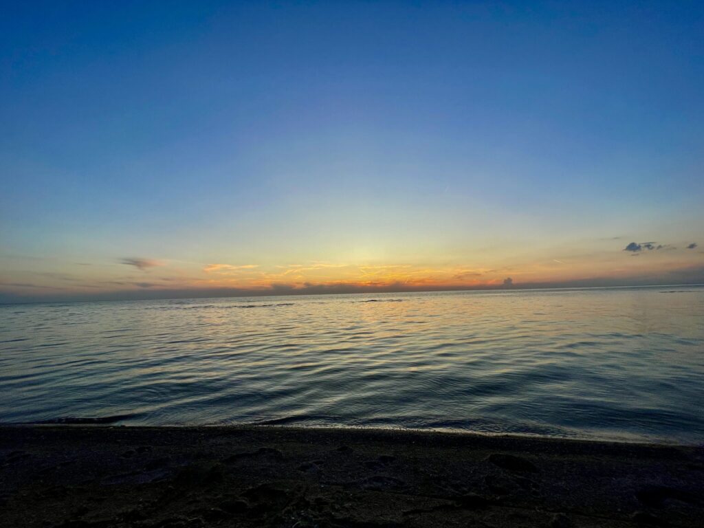 Calm Manitoba lake at sunset with golden light reflecting on the water, viewed from a dark sandy beach under a clear blue sky