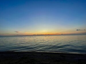 Calm Manitoba lake at sunset with golden light reflecting on the water, viewed from a dark sandy beach under a clear blue sky
