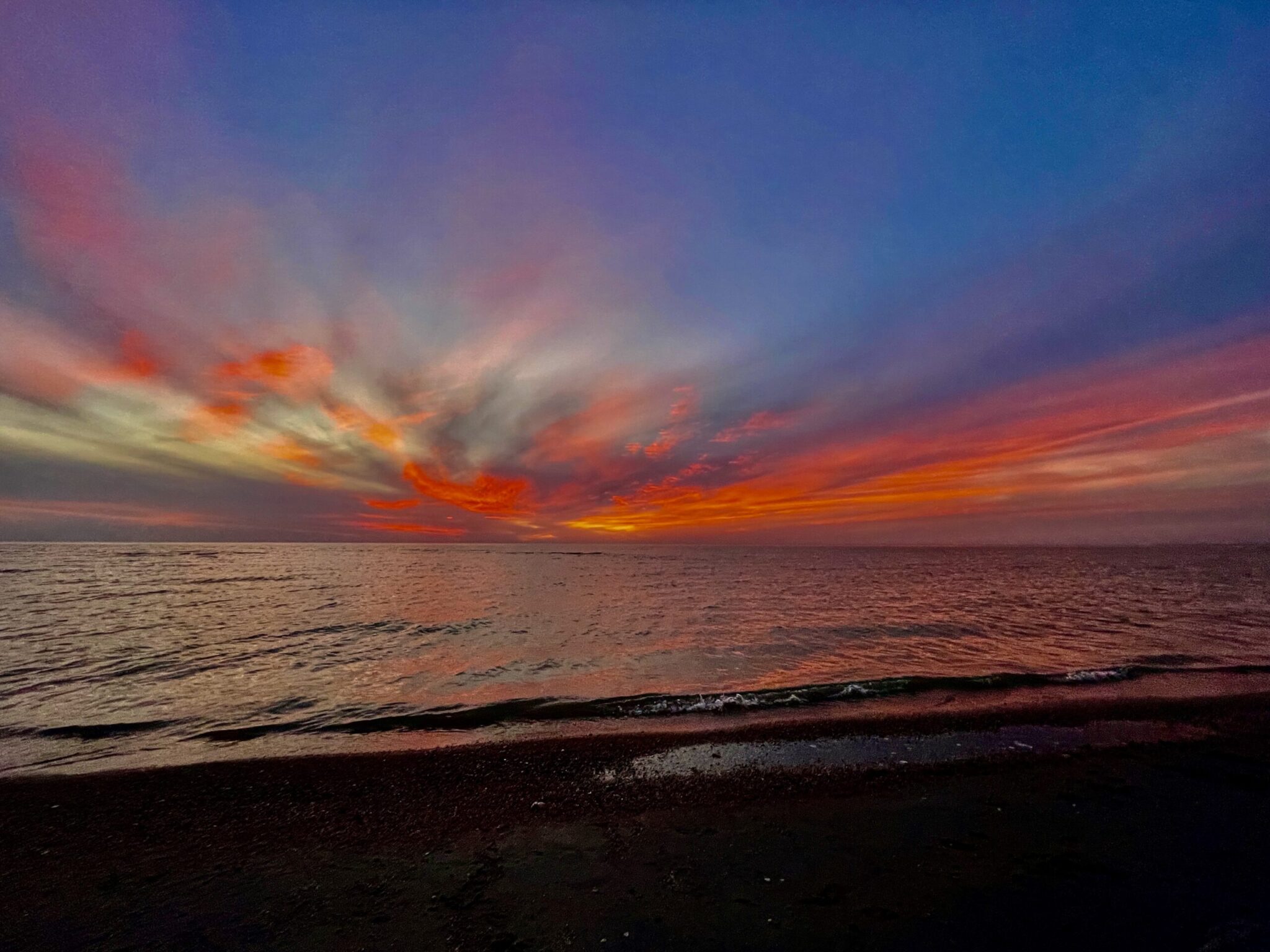 Vibrant sunset over a calm Manitoba lake with orange, red, and yellow clouds reflected in the water, dark sand beach in the foreground