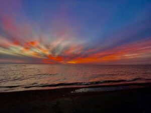 Vibrant sunset over a calm Manitoba lake with orange, red, and yellow clouds reflected in the water, dark sand beach in the foreground