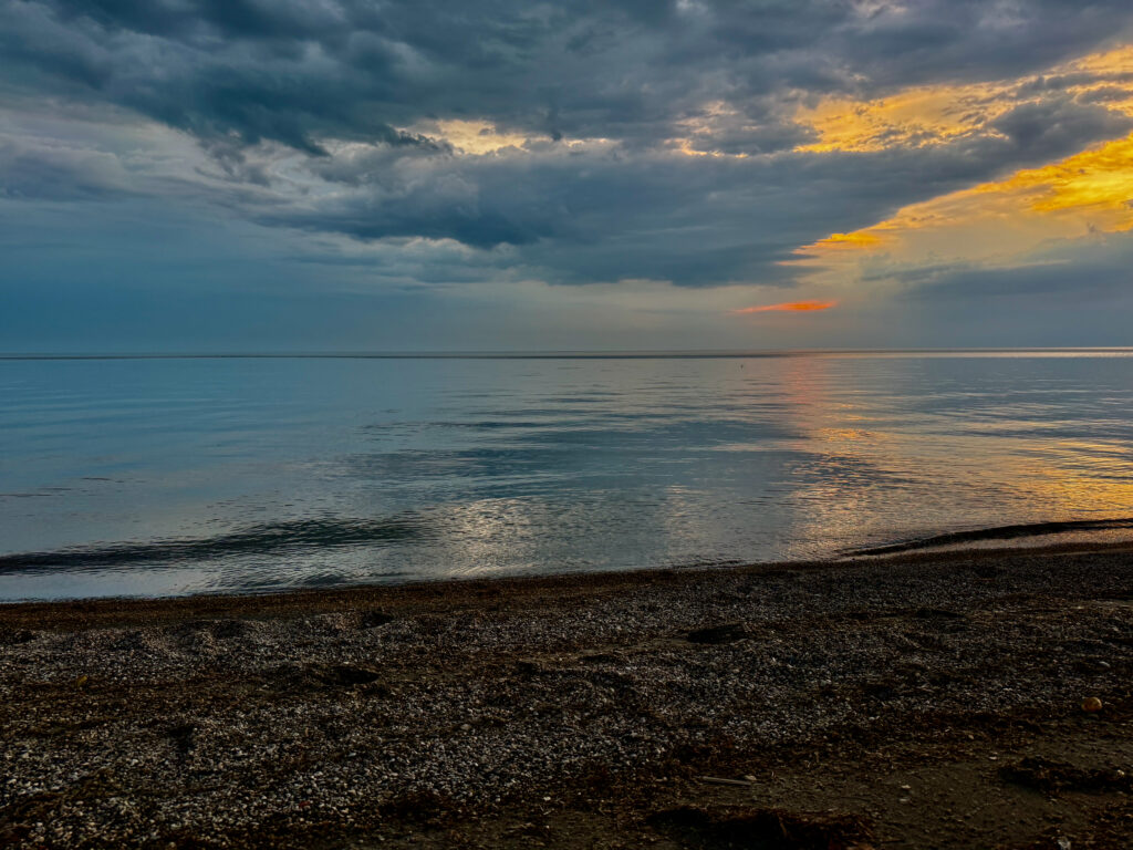 Calm beach at sunset with dark clouds overhead, golden light reflecting on still water, and a pebbled shoreline in the foreground