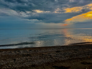 Calm beach at sunset with dark clouds overhead, golden light reflecting on still water, and a pebbled shoreline in the foreground