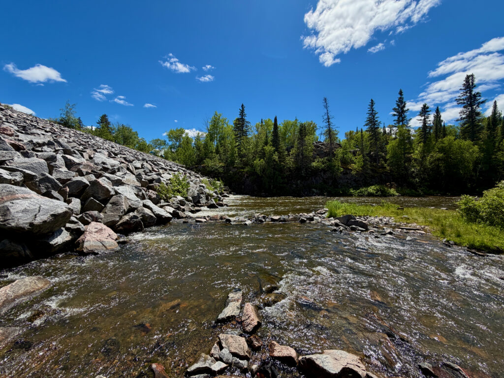 Rocky riverbank with flowing water, coniferous forest, and blue sky with white clouds on a clear summer day in a boreal landscape