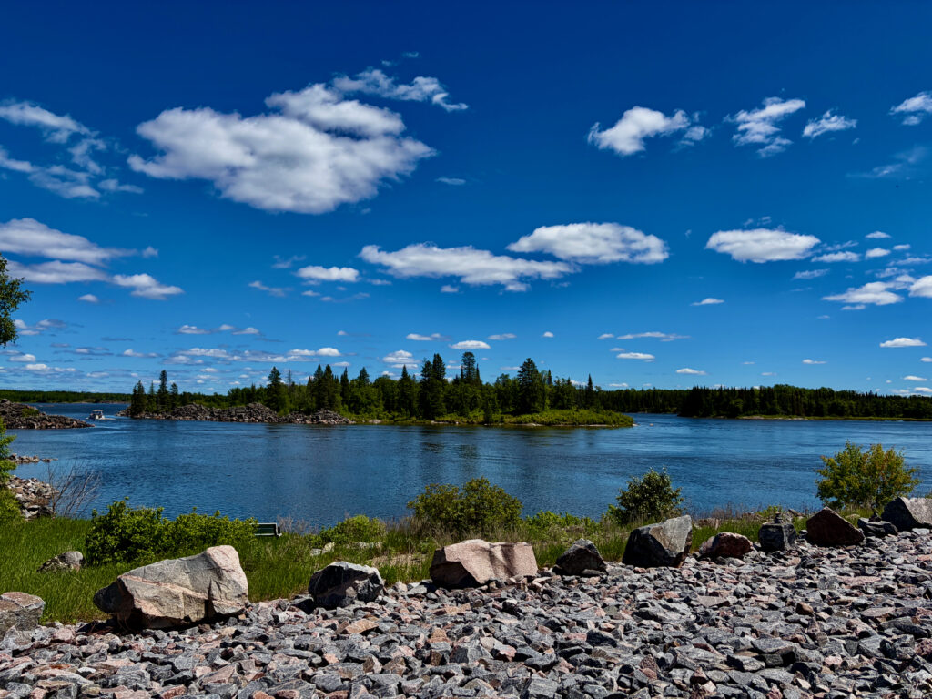 Rocky shoreline with blue water, forested islands, and clear sky with white clouds