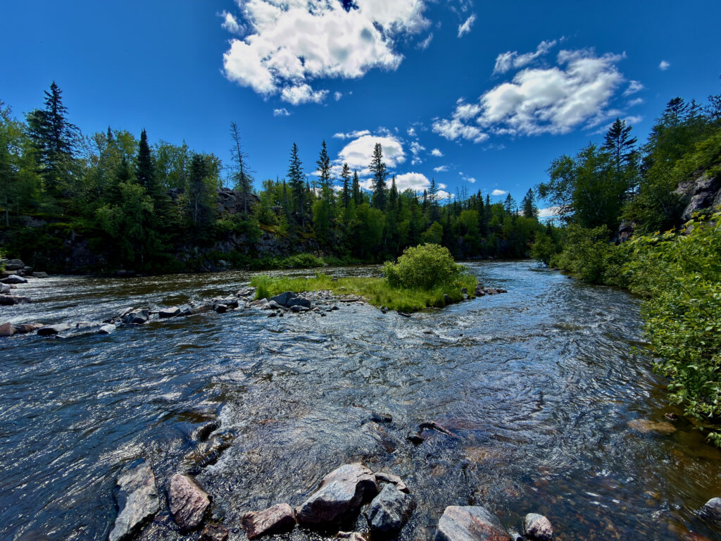River flowing through a boreal forest landscape with rocky shores, coniferous trees, and a clear blue sky with white clouds