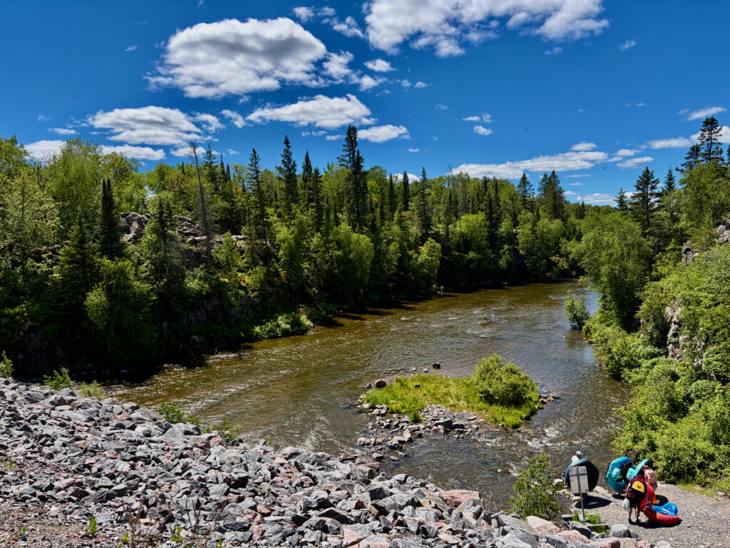 River flowing through boreal forest with rocky shoreline and canoeists preparing to launch in the foreground under a blue sky with white clouds