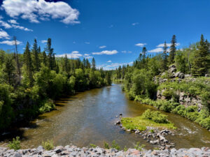 River flowing through a boreal forest landscape with coniferous and deciduous trees lining both banks, rocky outcrops visible on the right side, and clear blue sky with scattered clouds overhead